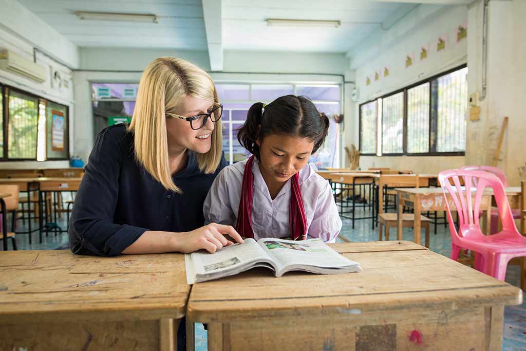 A blonde woman sits at a table in a classroom with a girl in w white shirt, reading a book together