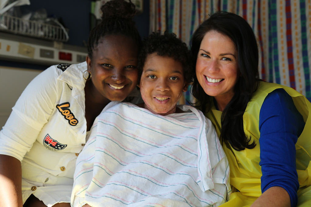 A boy in the hospital, covered by a blanket, sitting with his smiling sister and sponsor.