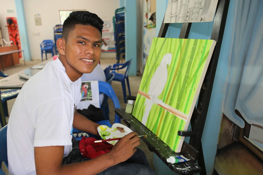 A young man sits infront of a paint canvas holding a paint brush and a paint palatte