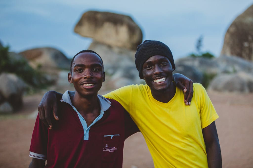 Twin brothers stand together with their arms around eachother's shoulders and smile.