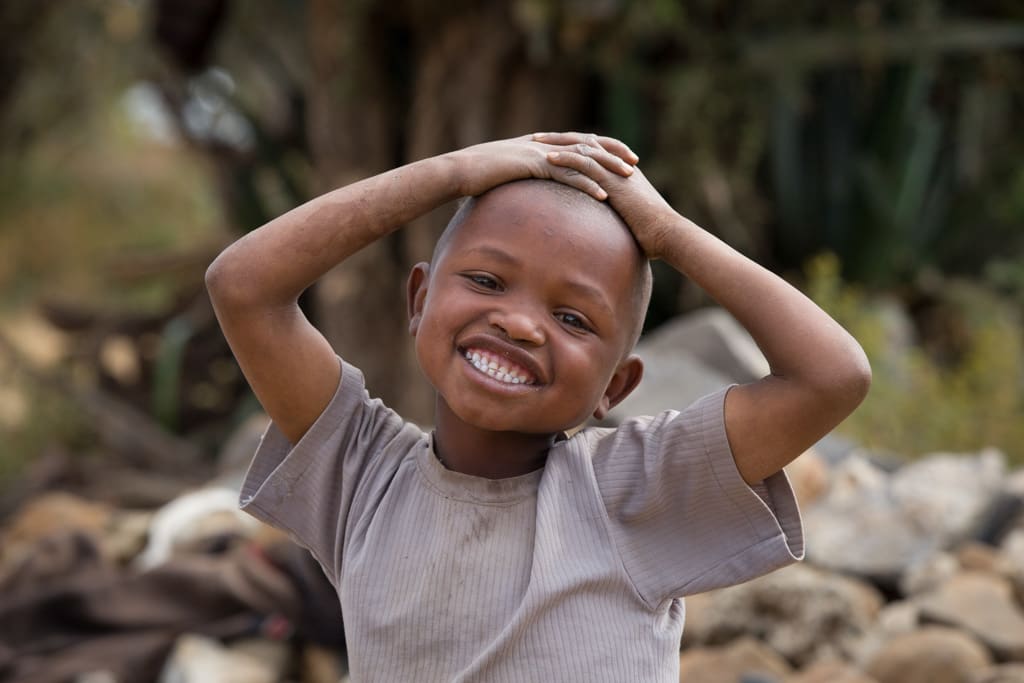 A boy, child, wearing a brown shirt, stares with happy, joy, smiling, expression on his face, outside with hands on top of his head.