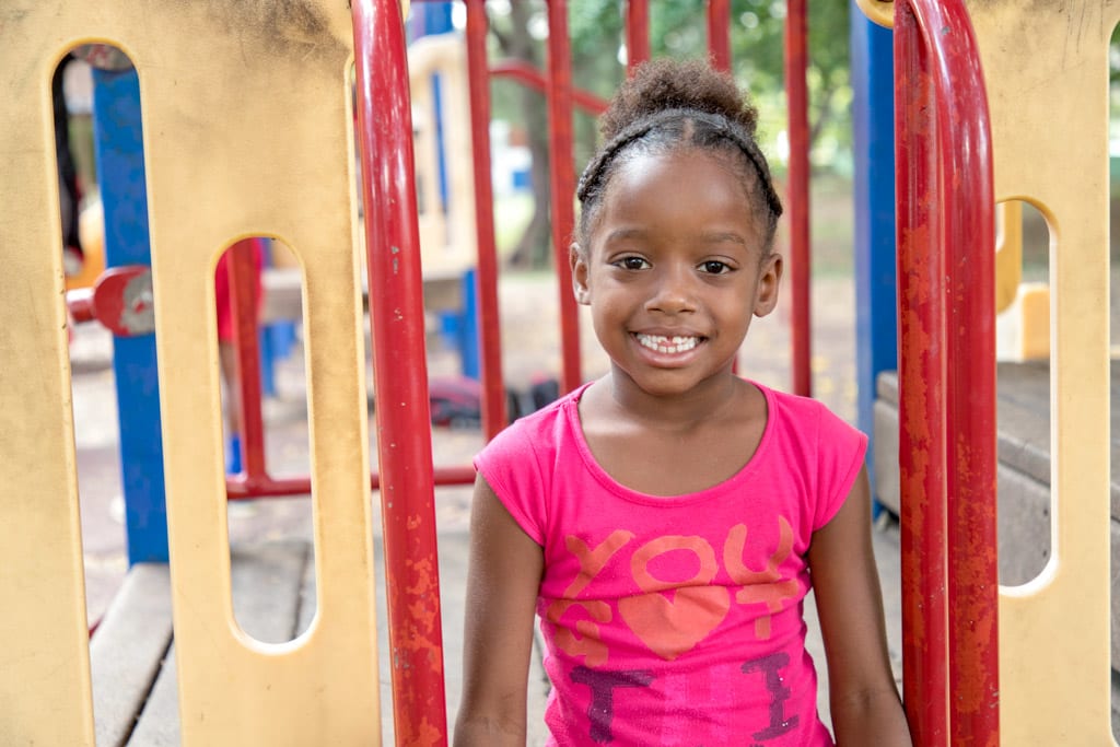 A girl in a pink t-shirt sits on a playground and smiles at the camera.