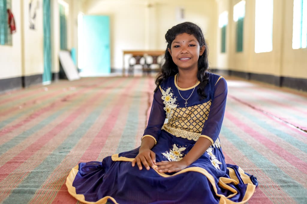 A girl sits on the floor in a large room. She wears an ornate dress and smiles.