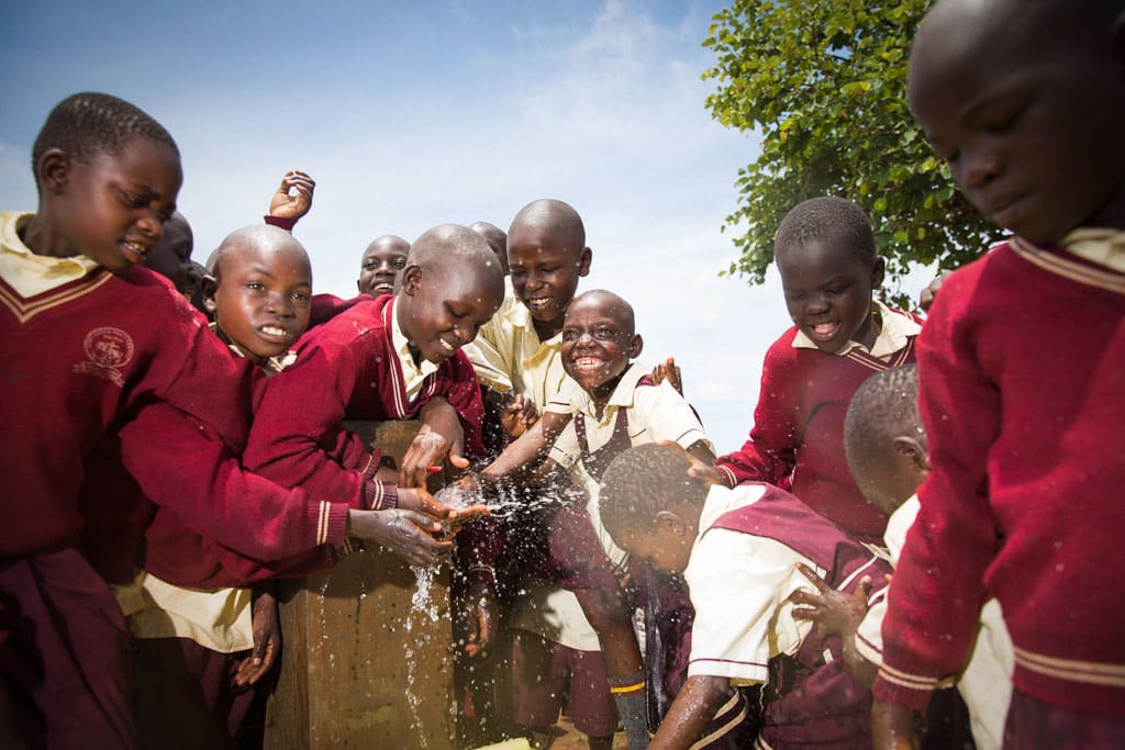 A group of children gather happily together, boys and girls, dressed in school center uniform clothes, red and white dresses or sweaters and shirts, as they are splashing water, clean safe drinkable sanitary pouring water from outside water source, outdoor water faucet.