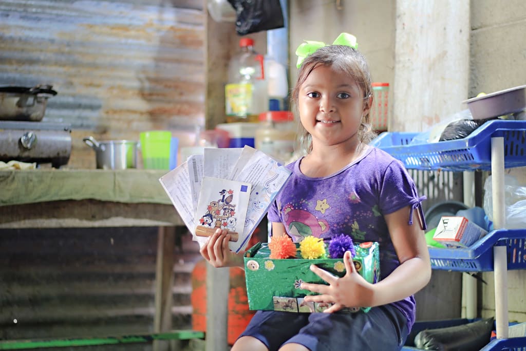 A little girl sits in a kitchen holding a shoe box full of letters and gifts from her sponors.