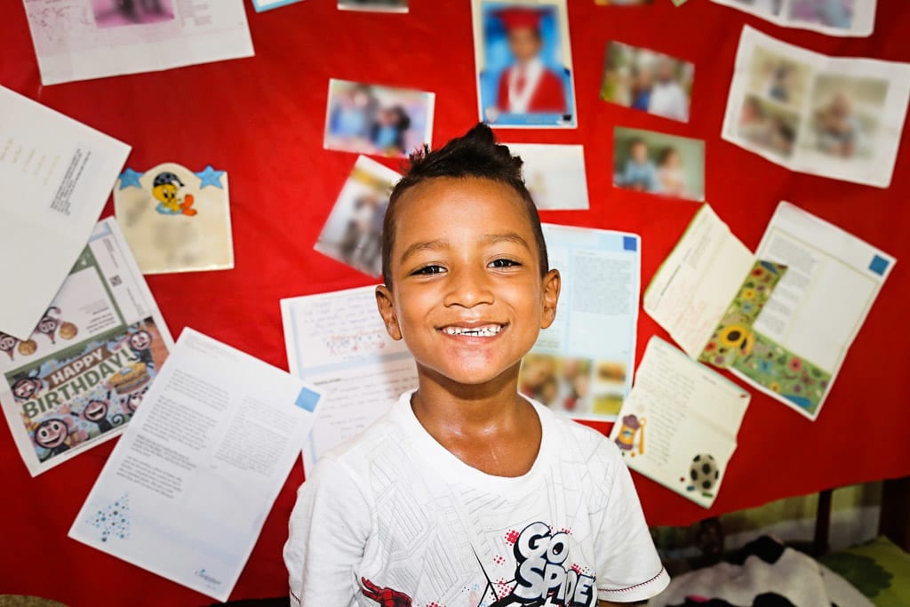 A boy sits infront of a red boad covered in pictures and letters. He smiles at the camera.