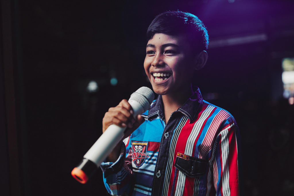 A happy boy, male child in a red, white, blue striped shirt holds a white microphone, sings, singing with stage lights behind him.