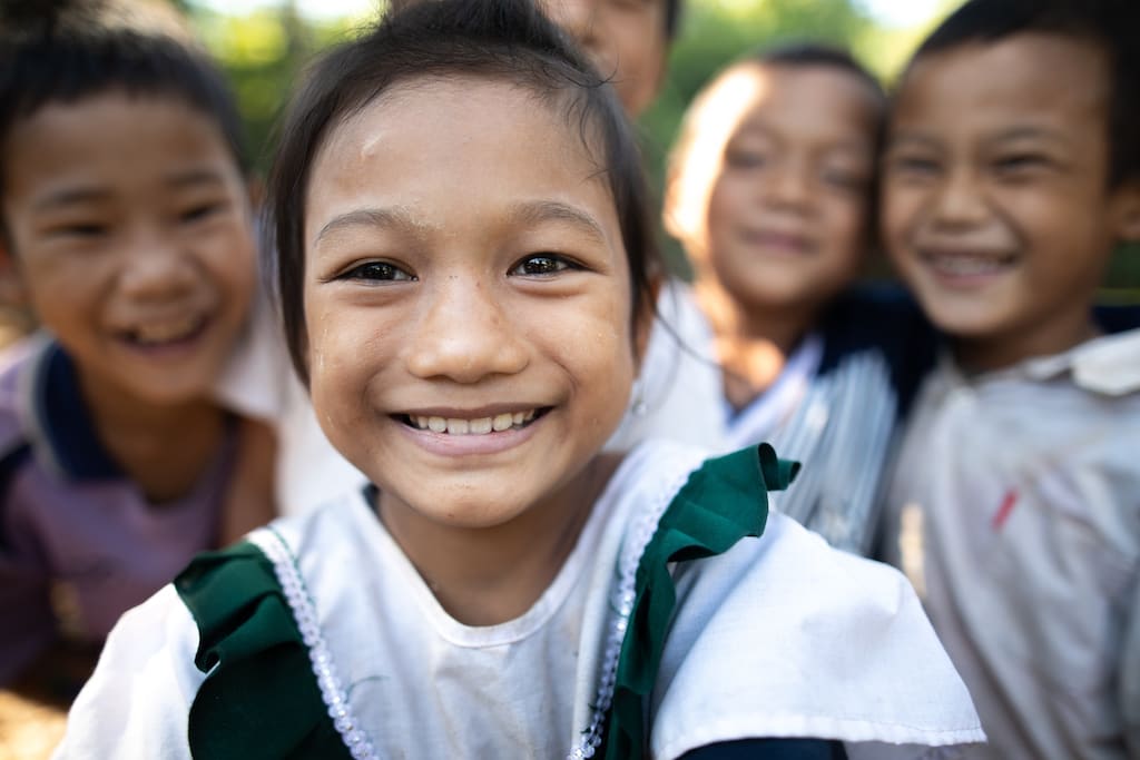 A Thai girl wearing a white and green shirt, with other children in the background.