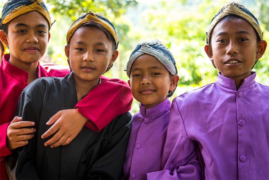 Four Indonesian boys stand together. They all wear ornate head scarves and silk, button up shirts.