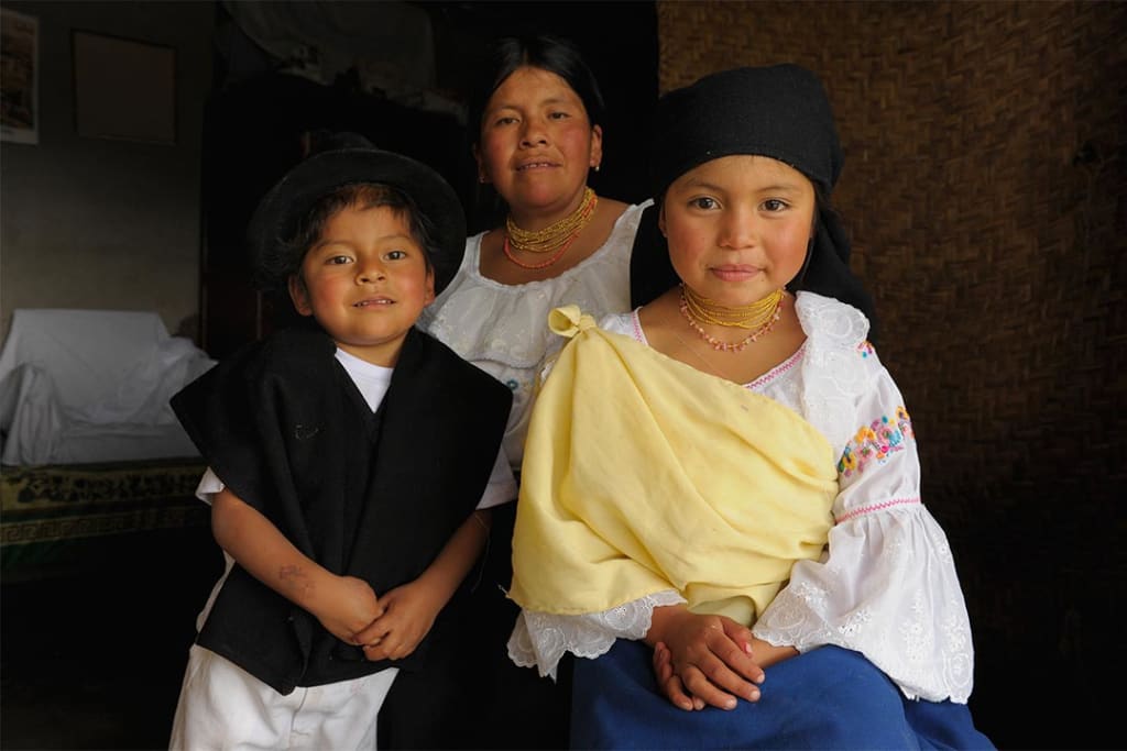 An Ecuadorian family of three sits and smiles at the camera. The daughter wears a black head scarf, an embroidered white blouse and a blue dress. Here mother is similarly attired. Her little brother where a traditional black hat as well as a black vest and white pants.