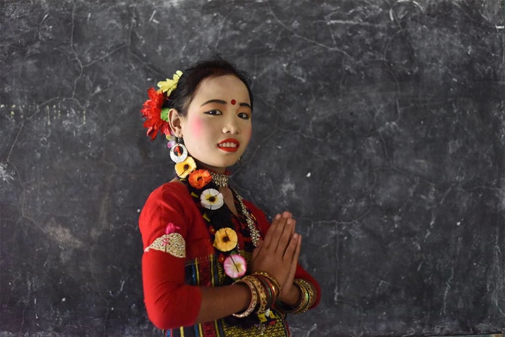 A young woman from Bangladesh where's traditional garb. She wears read lipstick, blush and eyeshadow and a red dot is painted on her forehead. She has flowers woven into here braided hair and bracelets cover her wrists. She wears an intricately embroidered red shirt.