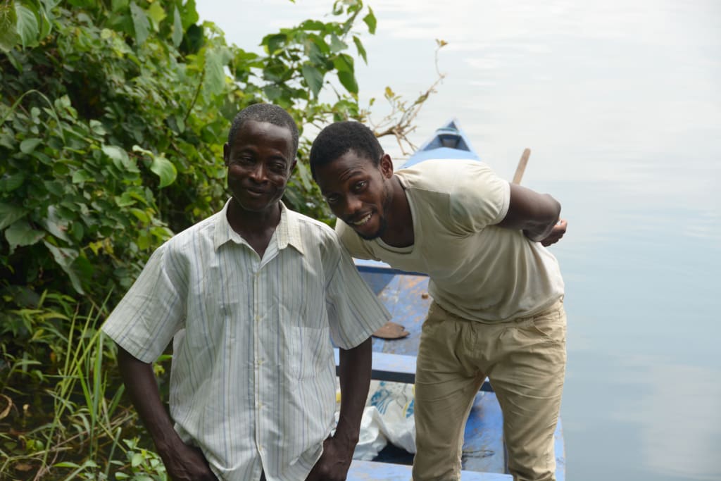 William Gasese with his friend and co worker Kofi, pose smiling together outside near a lake and boat