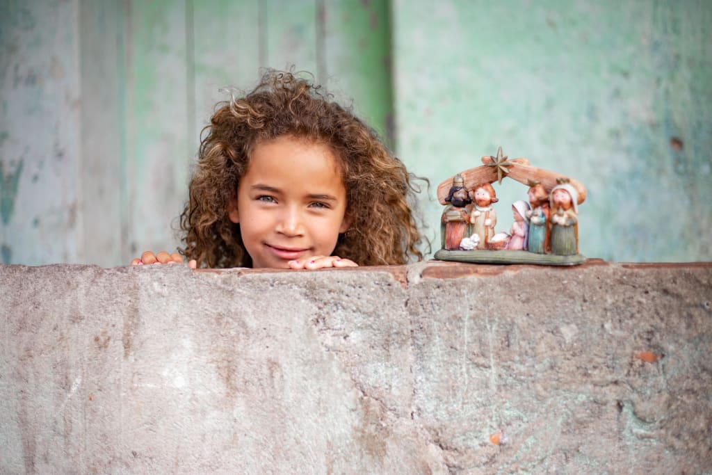 A girl with brown curly hair stands behind a concrete wall. She has a nativity set sitting beside her on the wall.