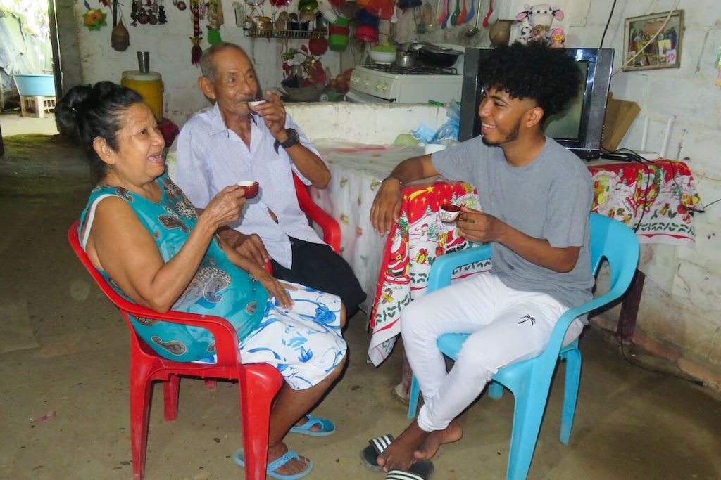 Oned is wearing a gray shirt with white pants. He is sitting inside his home and is having tea with his grandparents. They are all sitting in plastic chairs around a table.