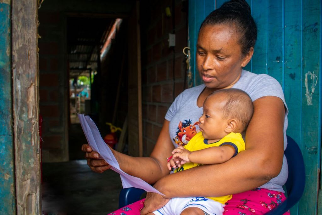 Bridis is wearing a gray shirt. She is holding her grandson, Deinner, who is wearing a yellow outfit, and is reading him a letter she wrote for him.