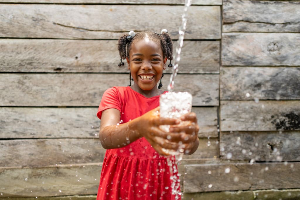 In Colombia, 9-year-old Ester is smiling as she holds a glass under a stream of clean, filtered water. The water is splashing in the air around her.