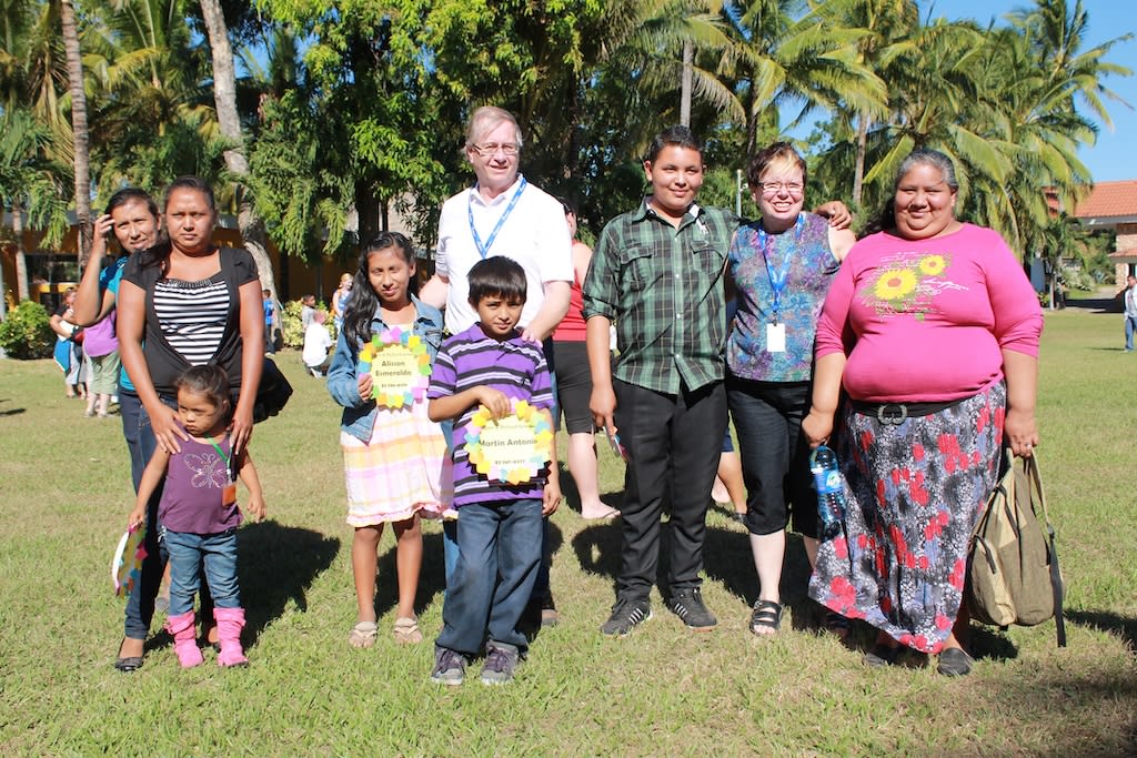 Lynn is pictured with 4 adults and 4 children. They are outside with palm trees behind them. They are standing on grass and smiling.