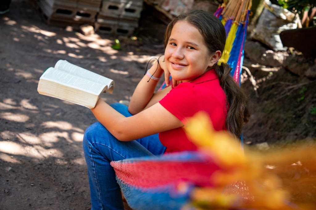 Greisy, a young girl in Honduras, is sitting on a hammock and holding her Bible.