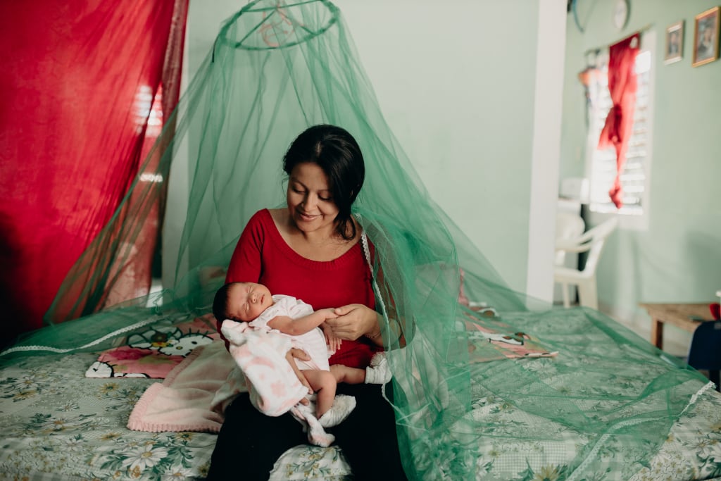 Brenda holds baby Samantha while sitting on a bed under a green mosquito net. It truly is a gift of health.