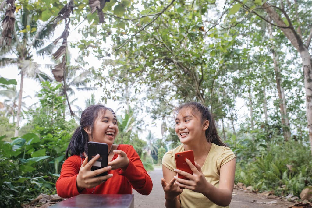 Two young girls are in a forest holding cell phones and smiling at each other.