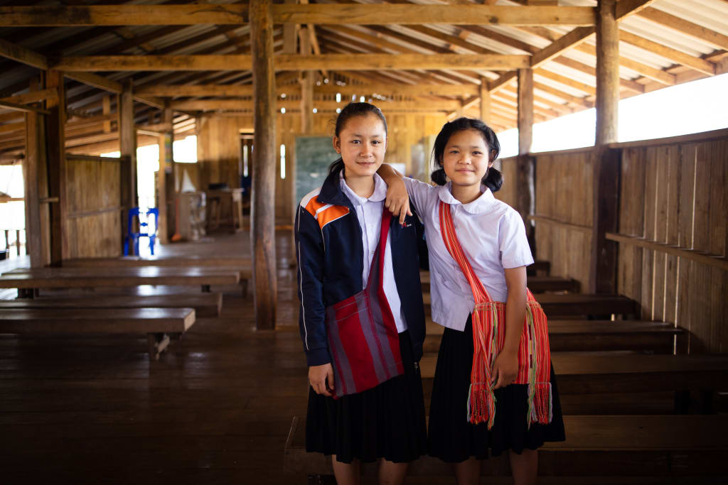 Two girls stand side by side in a classroom. They are wearing their uniforms and side bags.