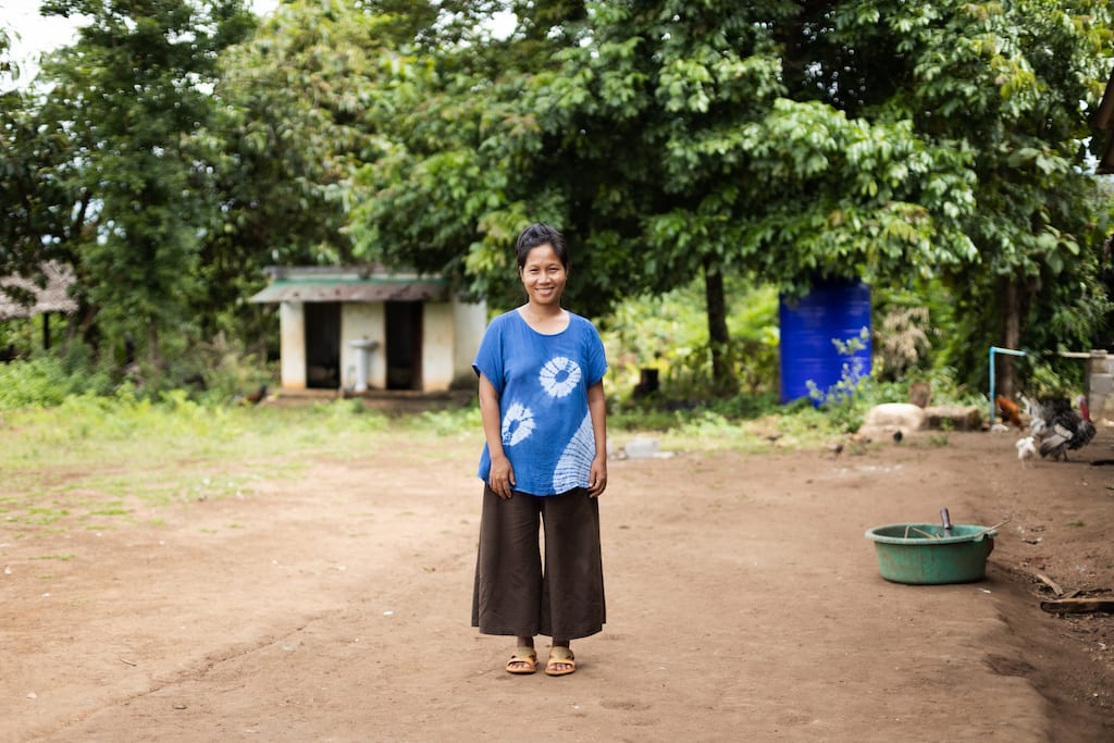 A woman stands on the first ground smiling. She has a tie-dye blue shirt and brown pants.