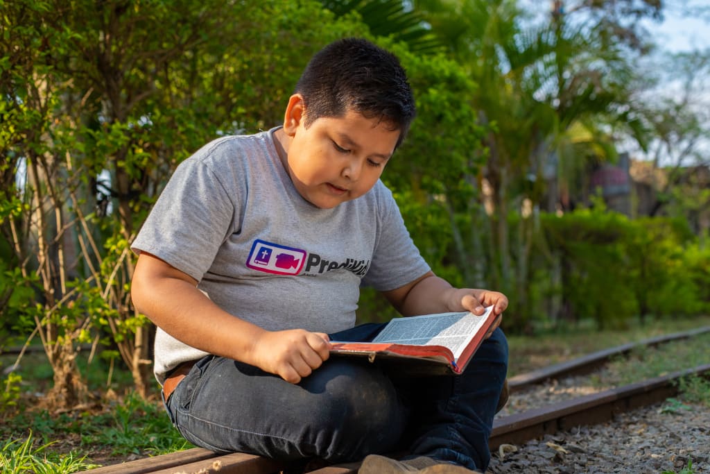 A boy sits on the ground reading a Bible in his lap.