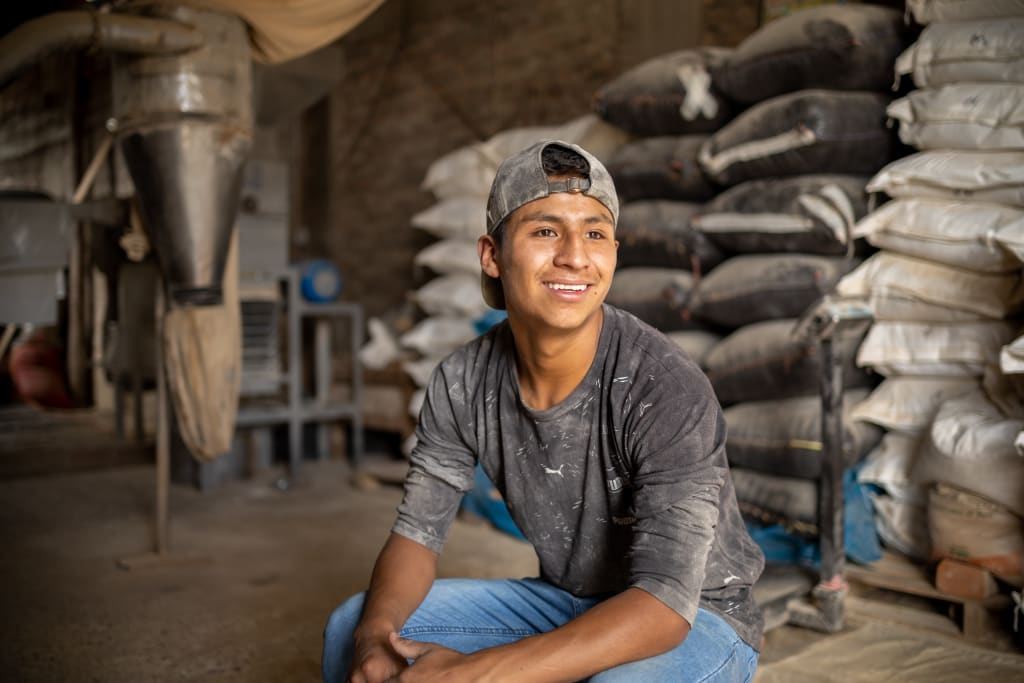 A young man with a cap and dusty clothes smiles in front of bags of grain at the warehouse for his small business. As he graduates the Compassion program, Angel has a secure small business.