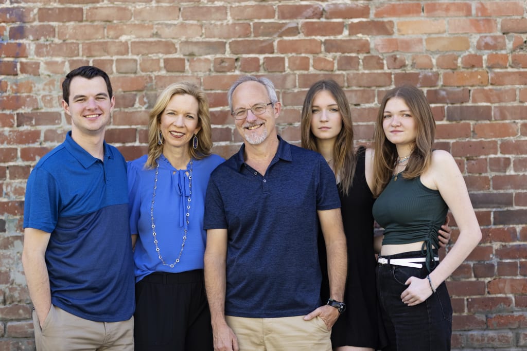 A photo of the Powell Family. Kara, Dave and their three young adult children smile against a brick wall.