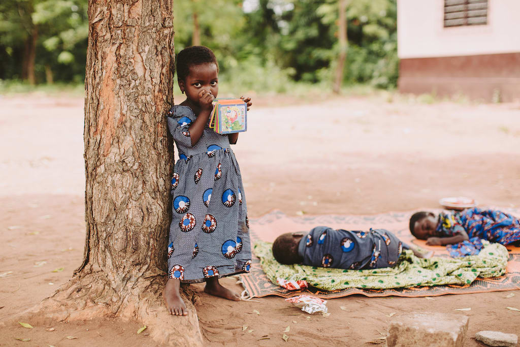 Little girl wears a blue patterned dress and holds a book. Babies are sleeping on blankets behind her.