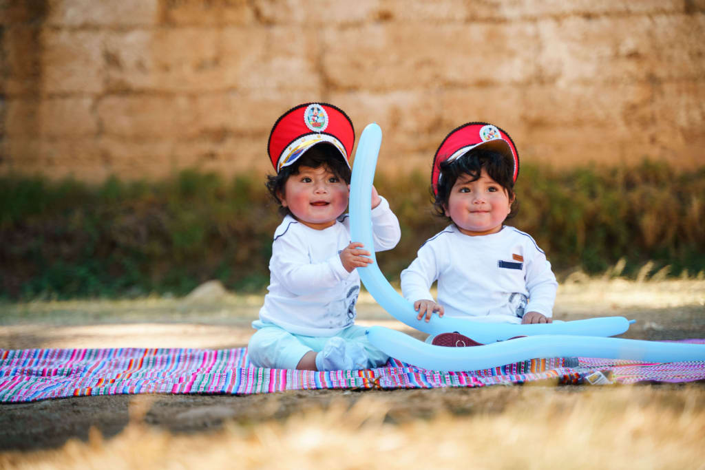 Two twins sit on a picnic blanket smiling holding balloons and wearing hats