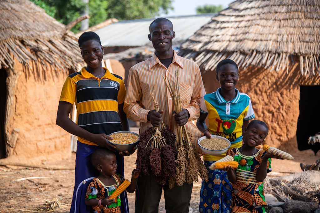 A family in Togo stands outside their home with crops they grew in a backyard farm. Backyard Farms are one of the gifts in the Gifts of Compassion gift guide.