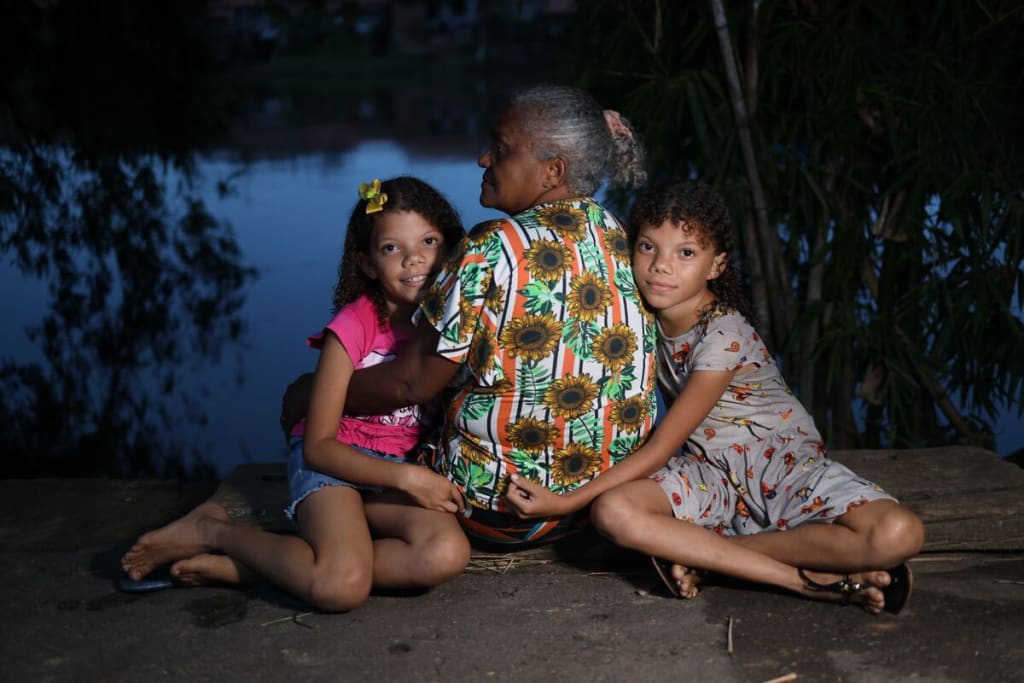 An older woman sits with her back to the camera and looks over her shoulder. Two young girls sit on either side of her and embrace her while looking at the camera.