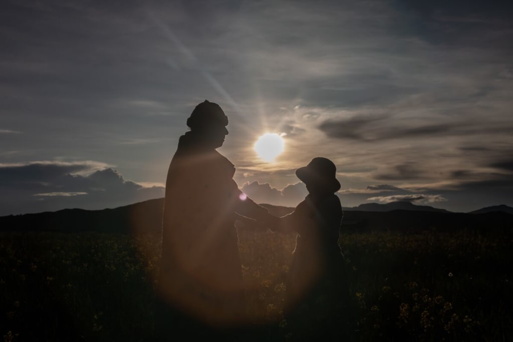 A woman and a girl hold hands in front of a sunset in a field.