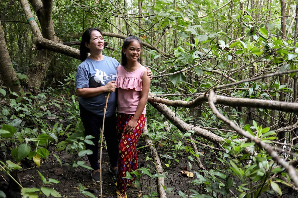 Kareen is wearing a pink shirt and red patterned pants. She is standing among the trees in the mangrove with Grace, the Project Director, wearing a blue shirt and holding a walking stick.