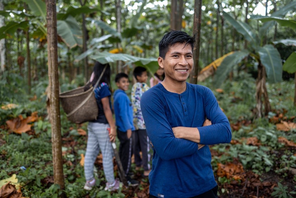 Gildo, in a blue shirt, is standing in the jungle with his arms crossed. The children are lined up behind him in the background.