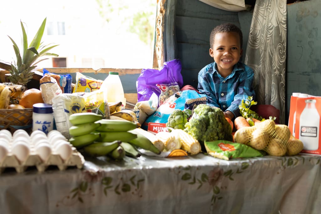 A boy sits smiling on a table full of vegetables and food items.