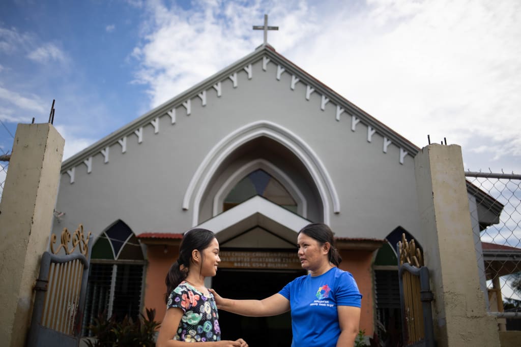 A girl in a floral top and a woman in a blue top stand in front of a church. They are facing each other and the woman has her hand on the girl's shoulder.