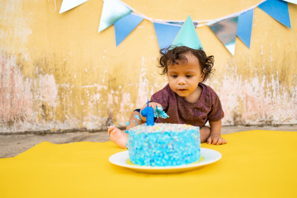 A boy wearing a blue birthday hat sits in front of a blue birthday cake.