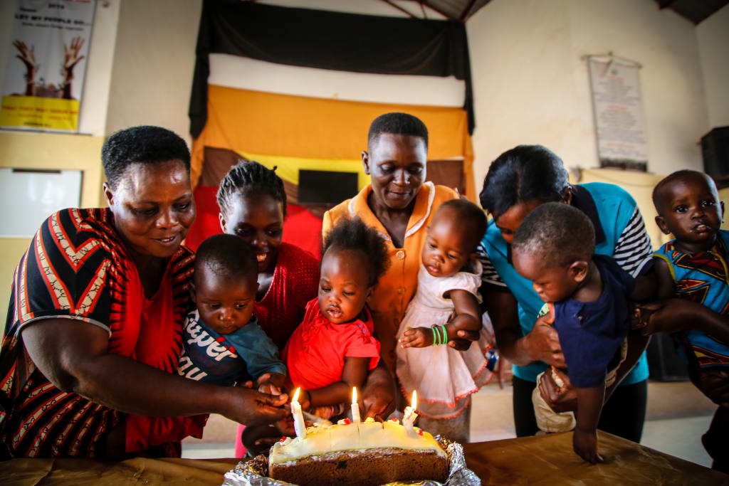 A group of mothers hold their one-year-old's up in front of a birthday cake. They are all smiling.