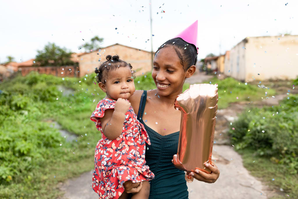 A mother wears a pink birthday hat and holds a "one" balloon in one hand and her daughter in the other.