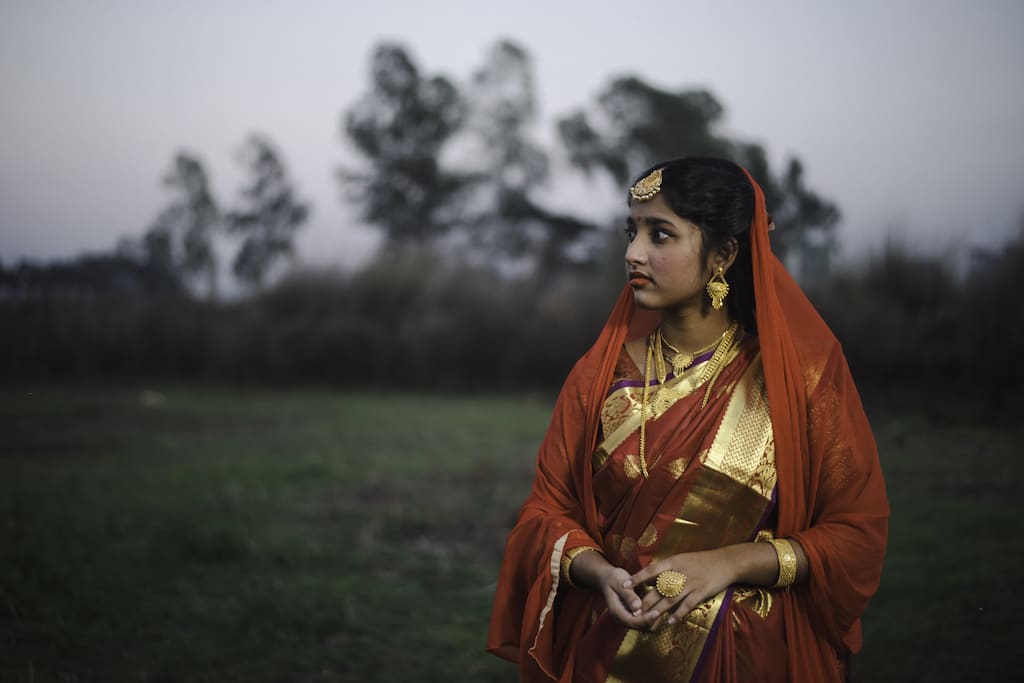 Tisha is wearing red and gold wedding clothing as part of a stage photoshoot protesting child marriage. She is standing outside and is looking off to the side.