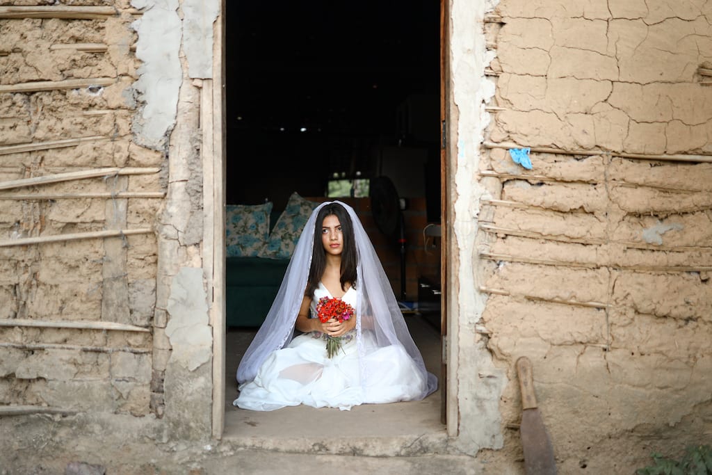 Yolane is wearing a white wedding dress and a veil for a staged photoshoot in protest of child marriage. She is sitting in the doorway of her home and is holding a bouquet of flowers.