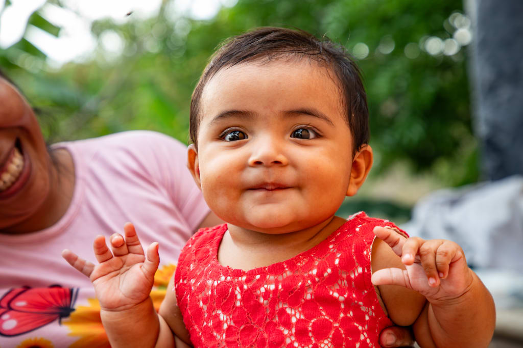 A baby wearing a red dress holds her hands up and smiles.