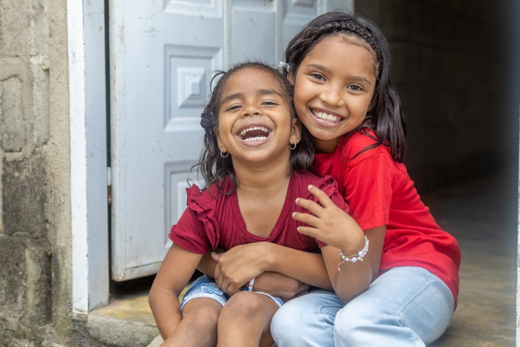 Two young girls in red shirts smile and hug each other while sitting on a front stoop.