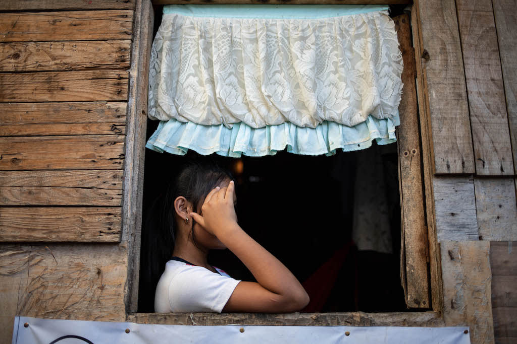 Girl holds her hand in front of her eyes. She is in the window sill with a curtain above her.