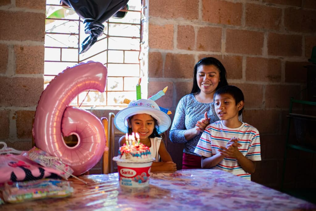A family celebrates a birthday and stands around a birthday cake with a balloon six