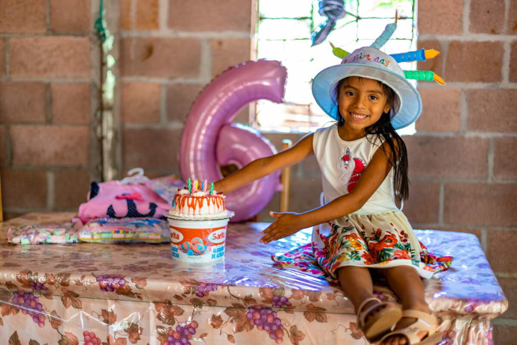 A girl sits with a birthday cake and a balloon six and smiles