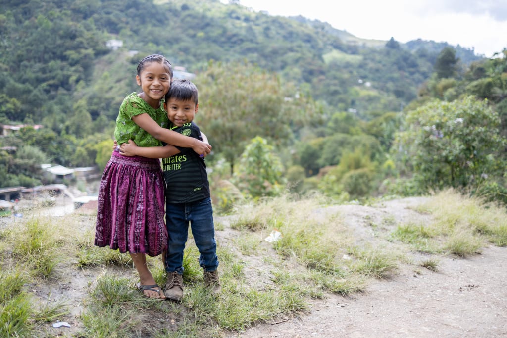 A young girl and boy stand in front of a hill and hug each other.