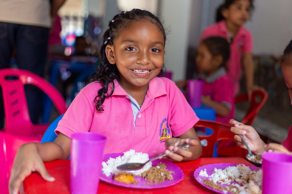 Girl wearing a pink shirt smiles while eating her dinner in the kitchen